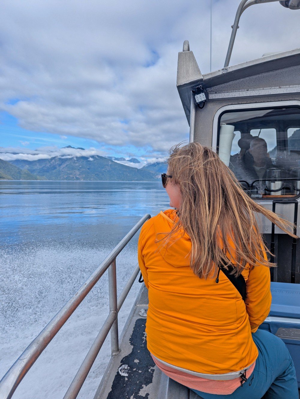 Back view of Gemma in an orange jacket sitting on side of boat looking out to ocean and mountain views in Bute Inlet