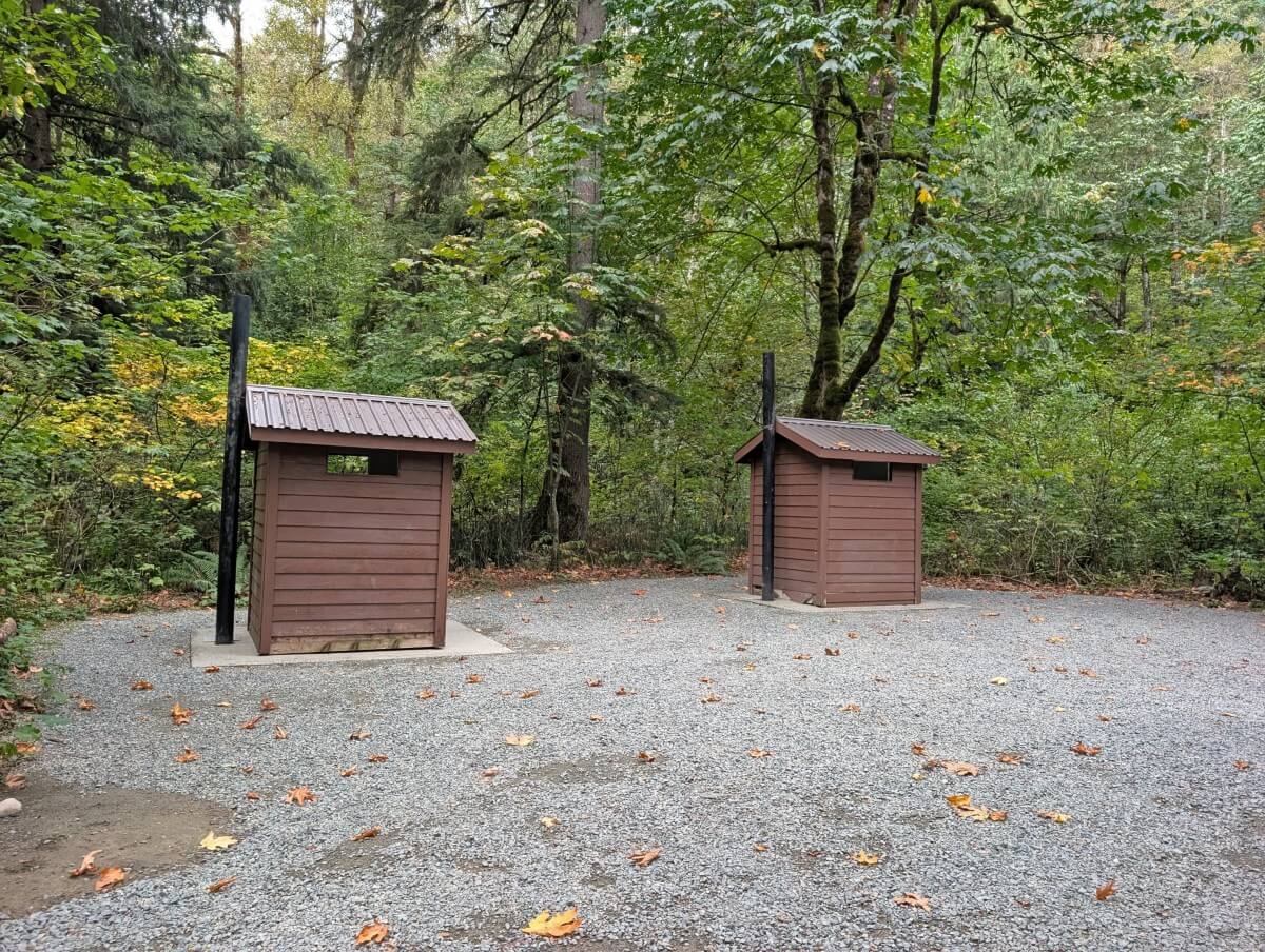 Two wooden outhouses in gravel area, backdropped by forest
