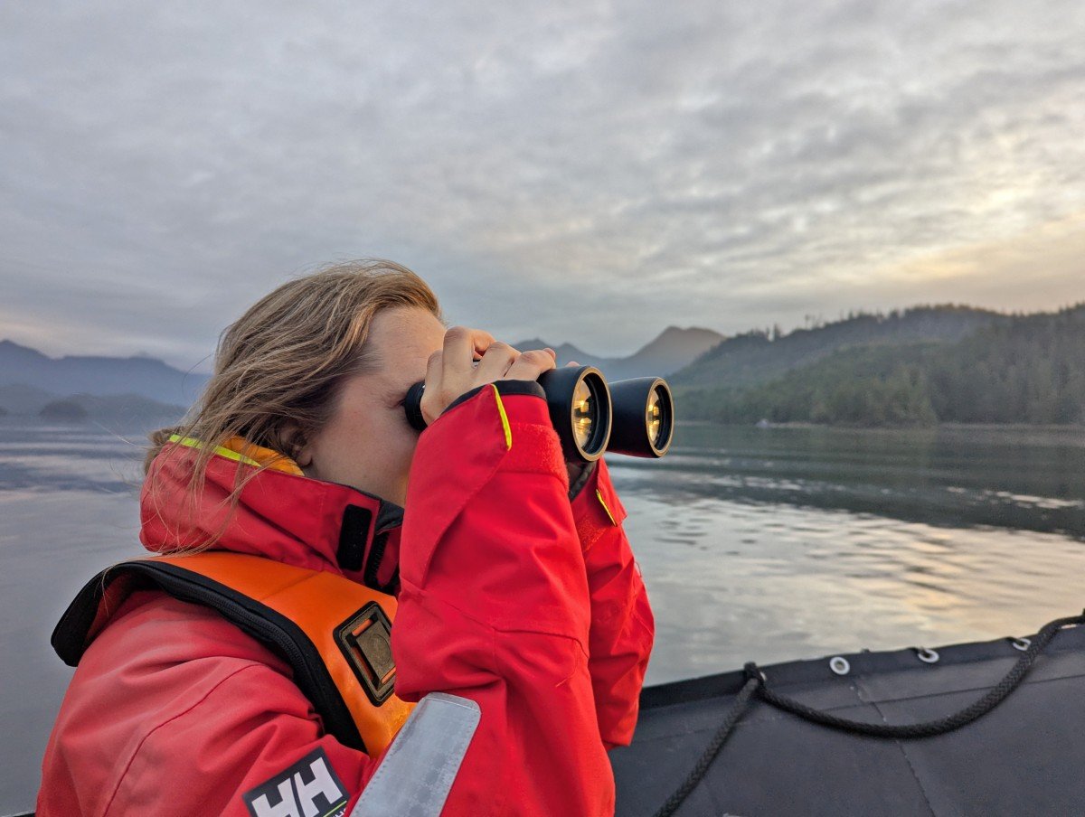 Close up of Gemma in red sailing jacket using binoculars to look for bears along the coast of Vancouver Island. There are layers of mountains and forest in the background