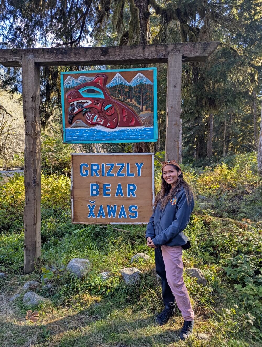 A Homalco guide (Kaylen) stands in front of a Grizzly Bear sign with First Nation bear artwork