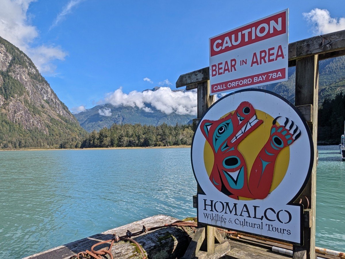 Close up of Homalco Wildlife and Cultural Tours sign featuring First Nation grizzly bear artwork. The sign above states 'Caution Bear in Area Call Orford Bay 78A.' The backdrop is forested mountains, with a scattering of clouds