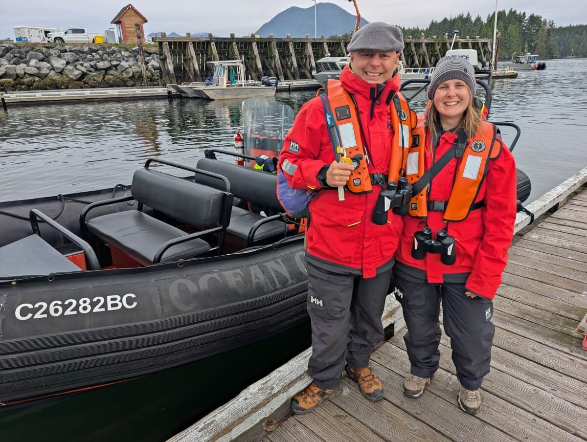 JR and Gemma stand in bright red sailing jackets and inflatable PFDs (deflated at the time) in front of black Zodiac boat in Tofino marina