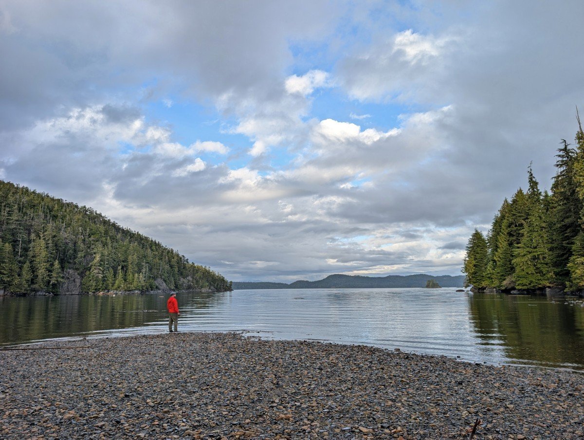JR stands on pebble beach in Bauza Cove near Telegraph Cove. The ocean is very calm and the edge of the cove is lined by forest