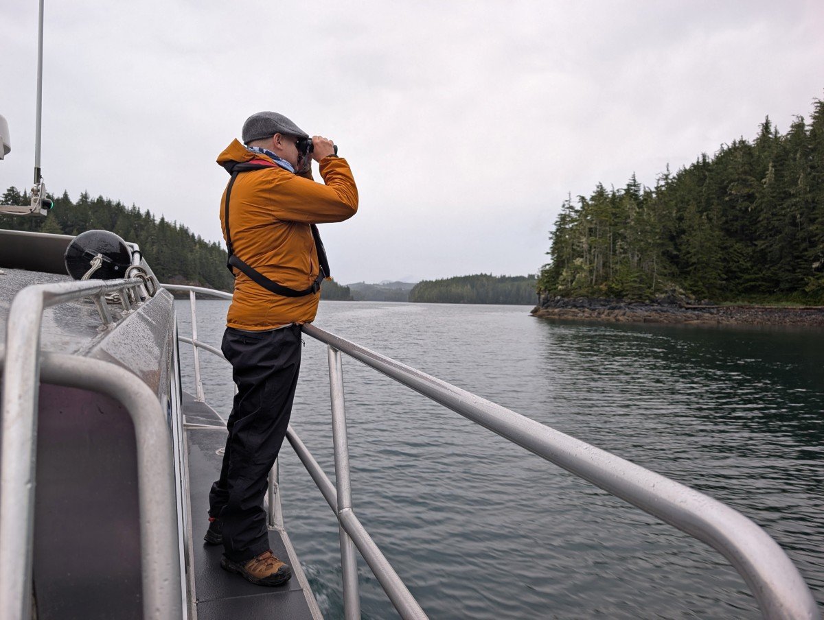 Sideview of JR standing on boat in Knight Inlet, using binoculars to watch a bear on Tide Rip grizzly bear watching tour