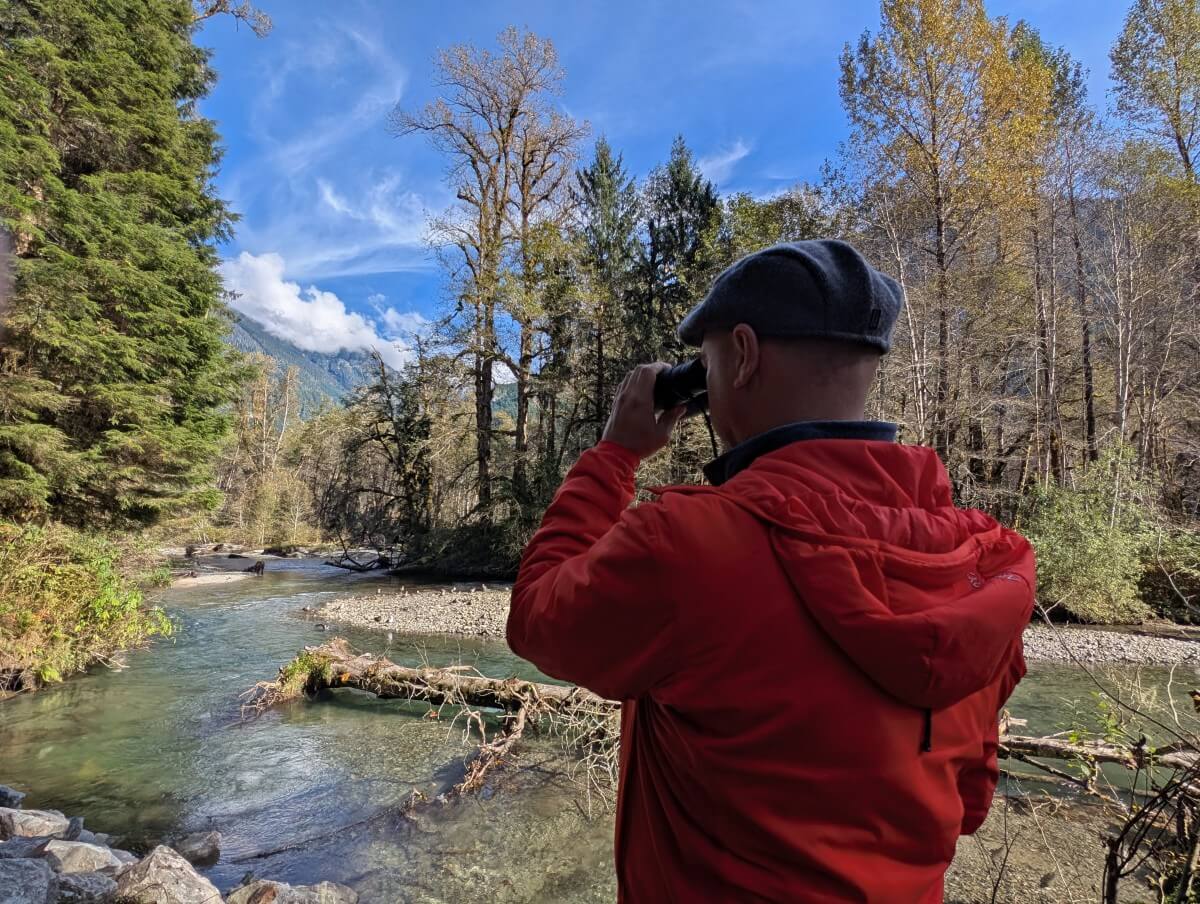 Back view of JR in a red jacket looking through binoculars at grizzly bear and cub in distance, next to the Orford River