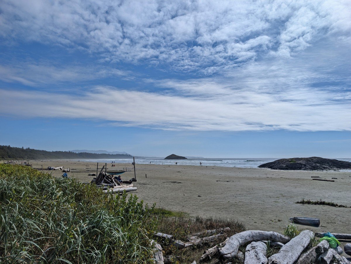 Long Beach view in Tofino, featuring a sandy beach stretching into the distance, driftwood in foreground and large rock to the right. The ocean is at low tide