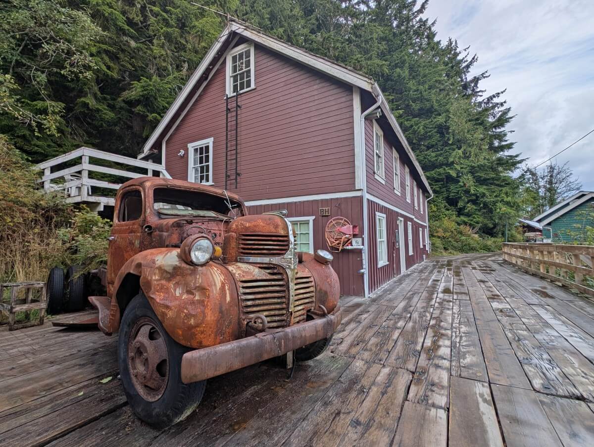 Side view of 1930s rusted truck parked on wooden boardwalk in Telegraph Cove, with two storey wooden brown painted building with forested background