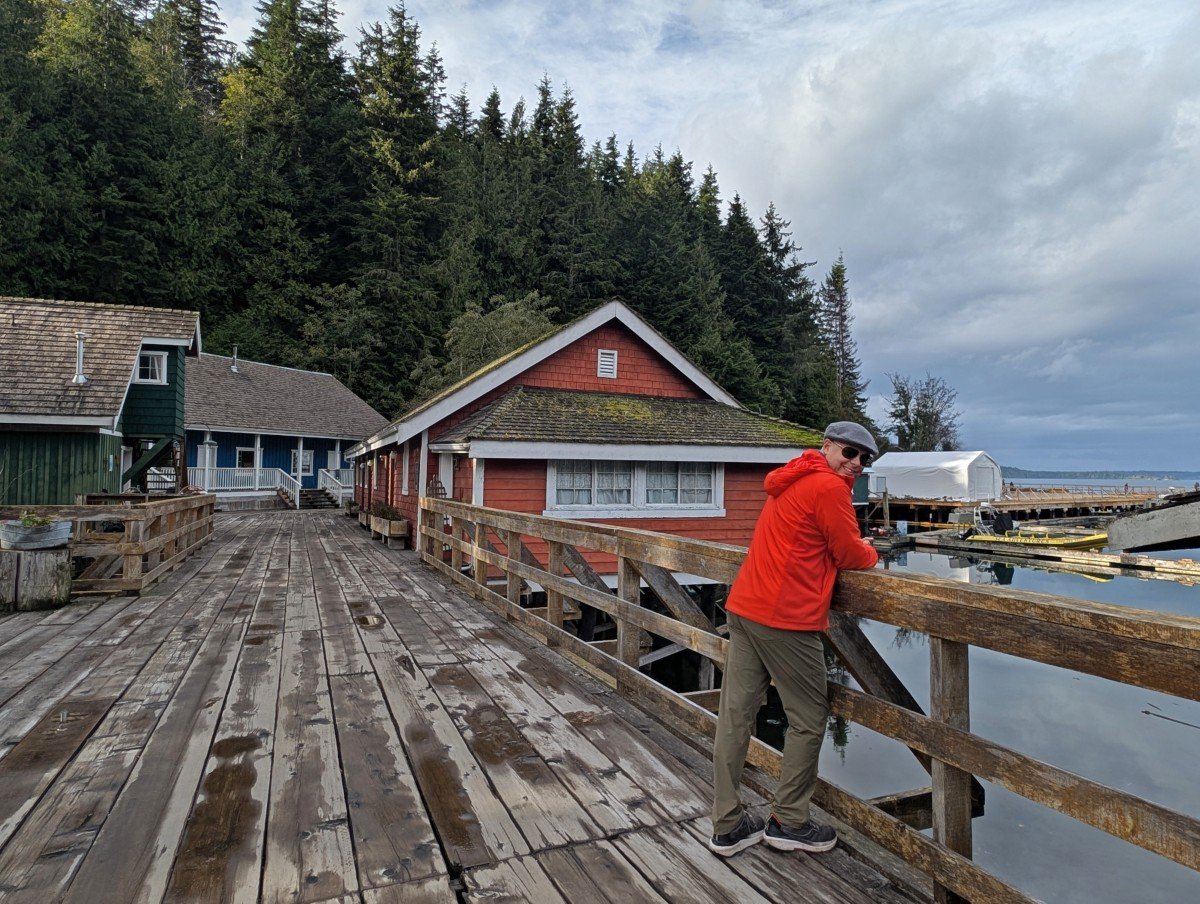 JR stands leaning on boardwalk fence in Telegraph Cove, with a backdrop of colourful wooden historical buildings
