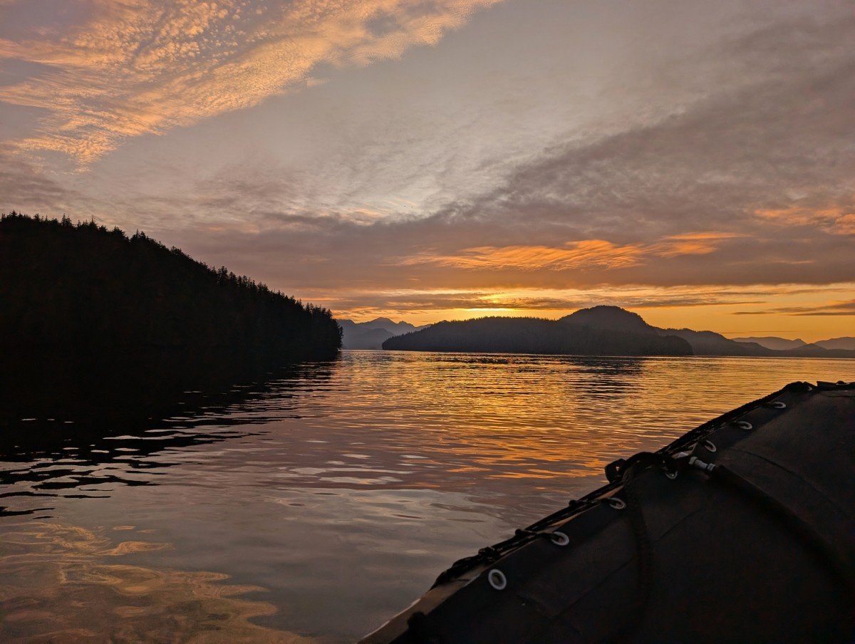 Sunrise view on Zodiac boat tour in Tofino, with orange colours behind layers of mountains and islands. Part of the Zodiac boat is visible to the right