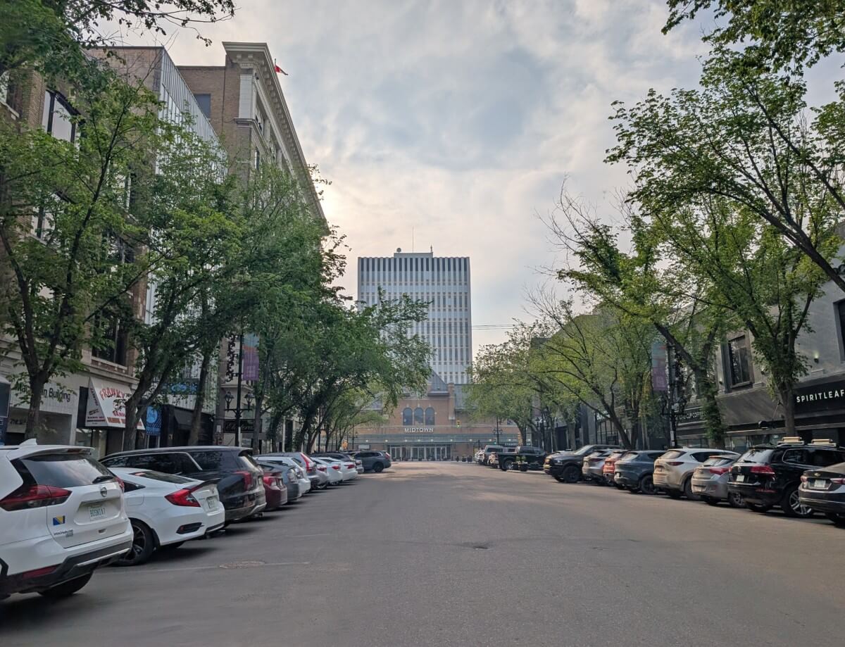 Road view of downtown Saskatoon featuring cars parked on both sides, old station (turned mall) ahead and trees lining each side