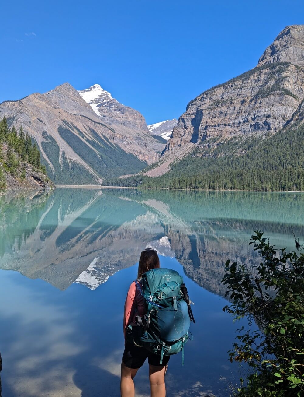 Back view of a hiker with a large backpack standing in front of calm Kinney Lake, a turquoise coloured lake reflecting mountain views