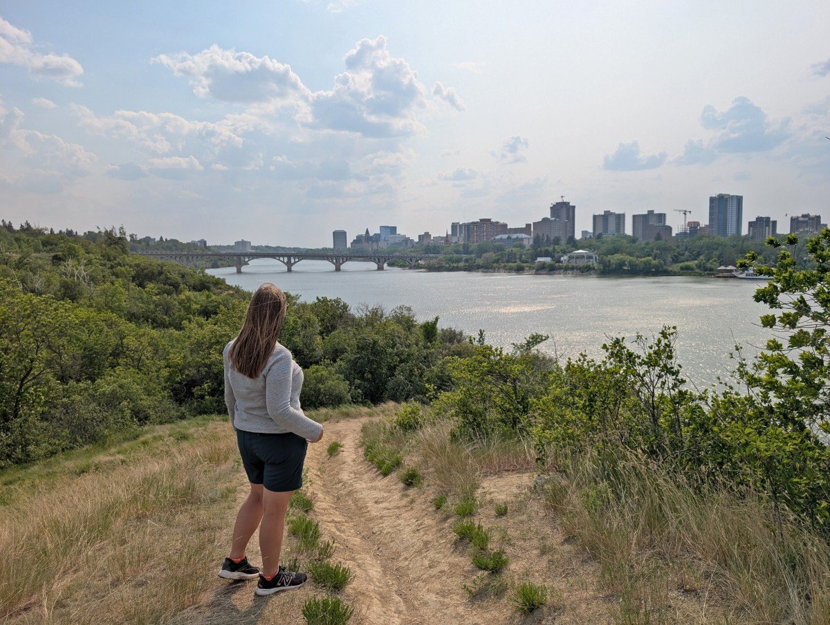 Back view of Gemma standing on hiking path above river, looking out to views of tall buildings in downtown Saskatoon across the water