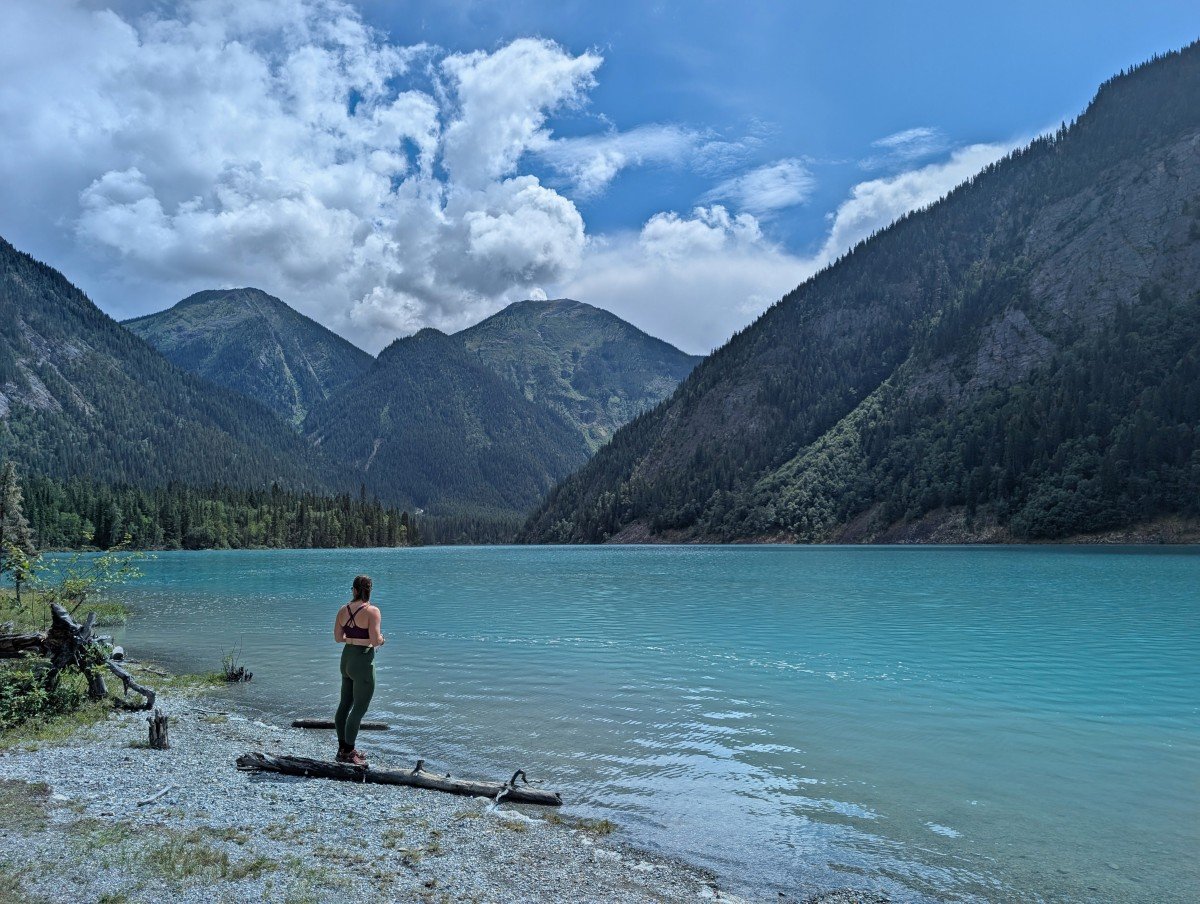 Back view of female hiker standing on the shores of Kinney Lake, a calm turquoise coloured lake backdropped by forested mountains