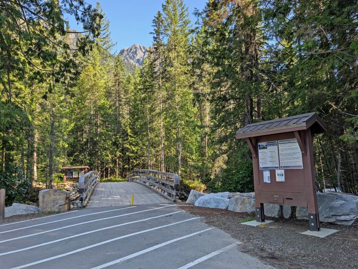 Kinney Lake trailhead, featuring wooden kiosk with signage and a bridge leading over the river, all backdropped by forest