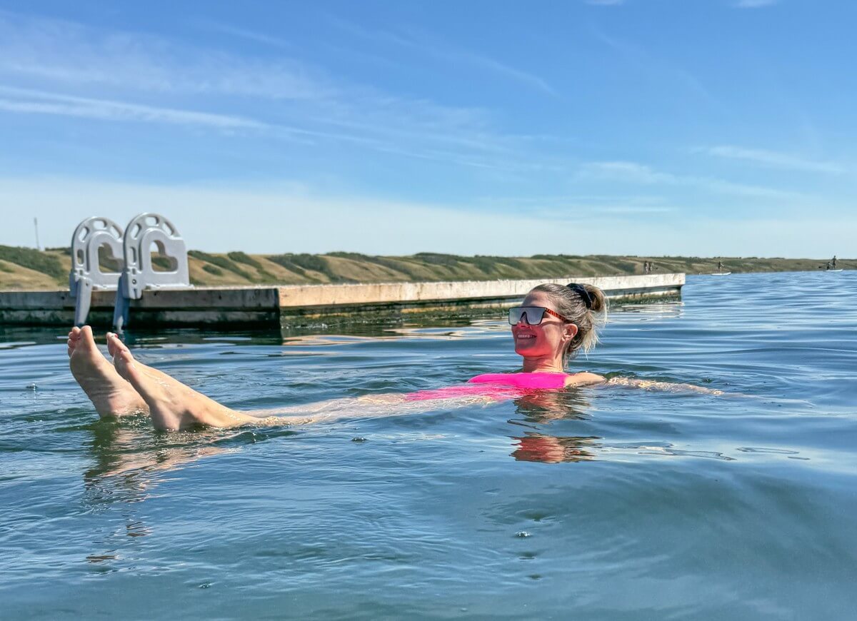Close up photo of woman in pink bathing suit floating in Manitou Lake, Saskatchewan, with floating dock behind