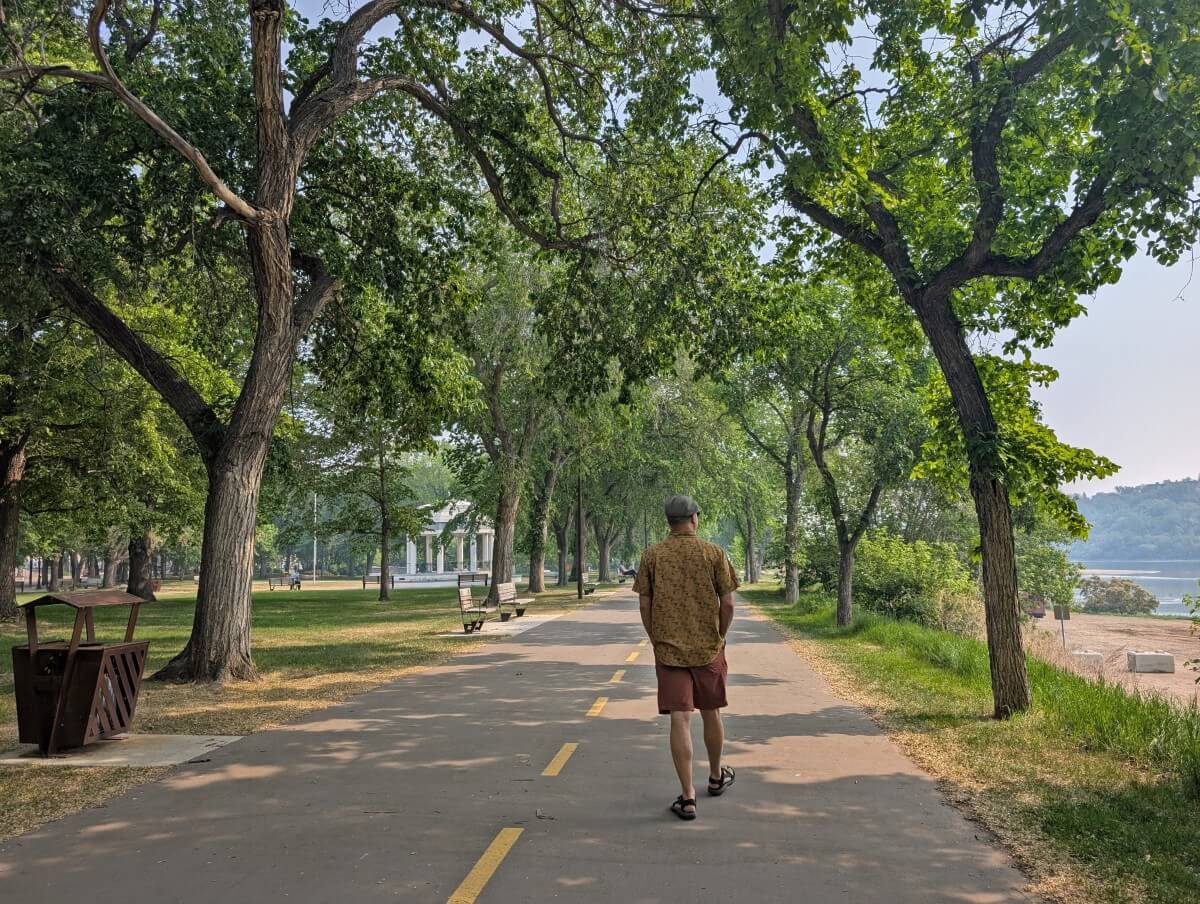 Back view of JR walking along a paved section of the Meewasin Trail in Saskatoon. The path is lined by trees