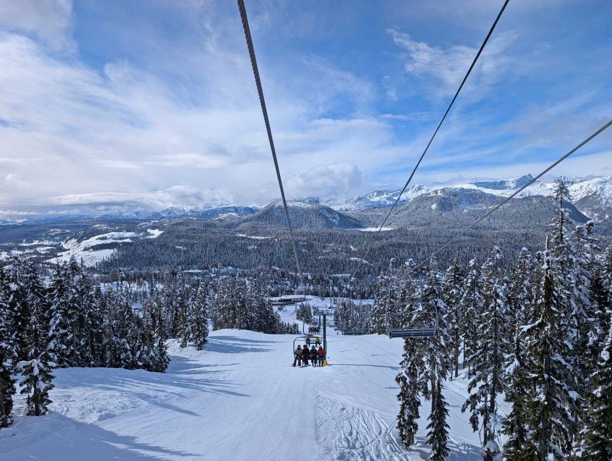 Chairlift view at Mount Washington looking back at chair behind, with views of snowy ski resort and snow capped mountains behind