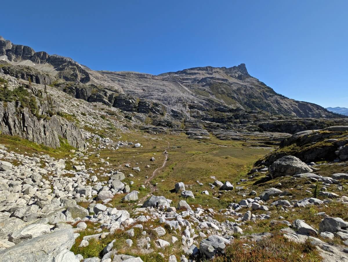Looking down on a faint dirt path running through alpine meadows cenery below, surrounded by rocky slopes and mountain peaks