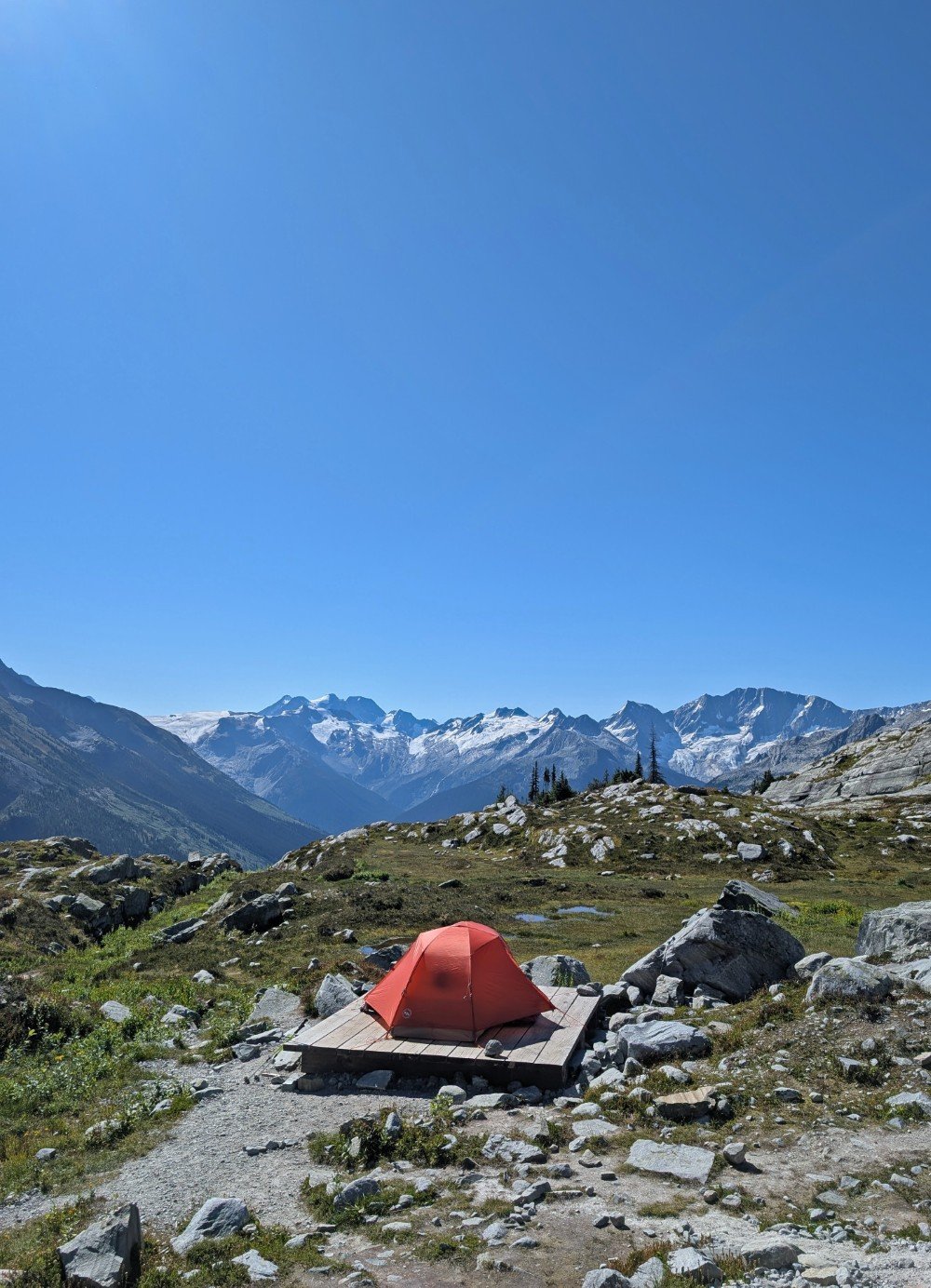 Orange set up tent on wooden platform in front of alpine meadows scenery, backdropped by snow capped mountains and glaciers