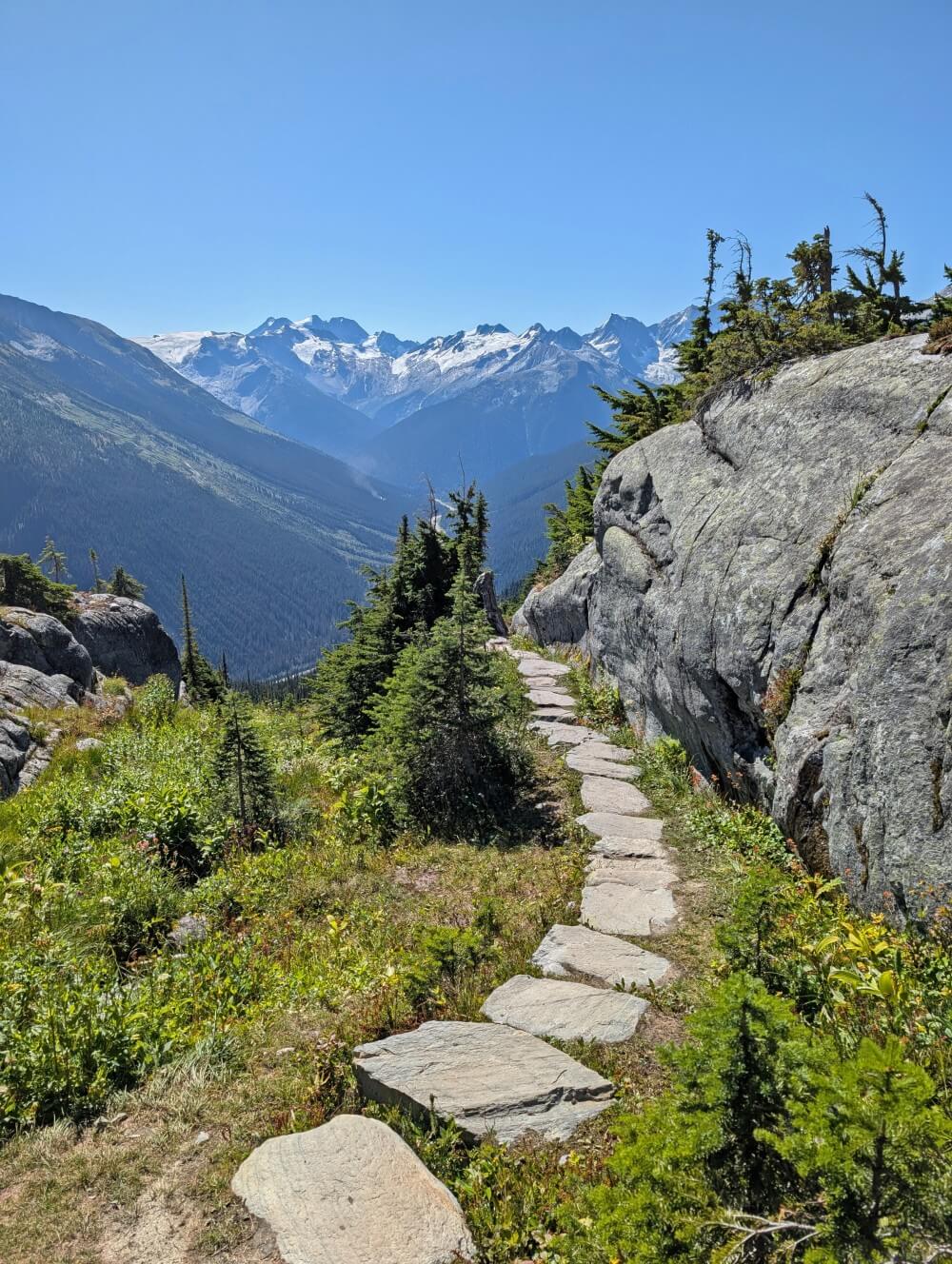 A series of rock steps lead the way along a large rock face to a downhill section of trail, backdropped by forest and mountain views
