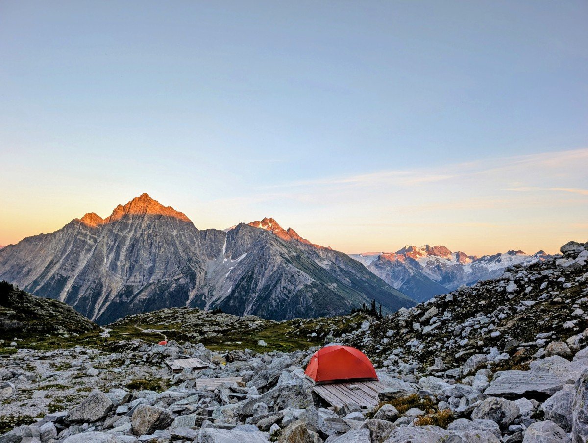 An orange tent sits in a boulder filled bowl backdropped by mountains at sunset - Hermit Meadows, Glacier National Park