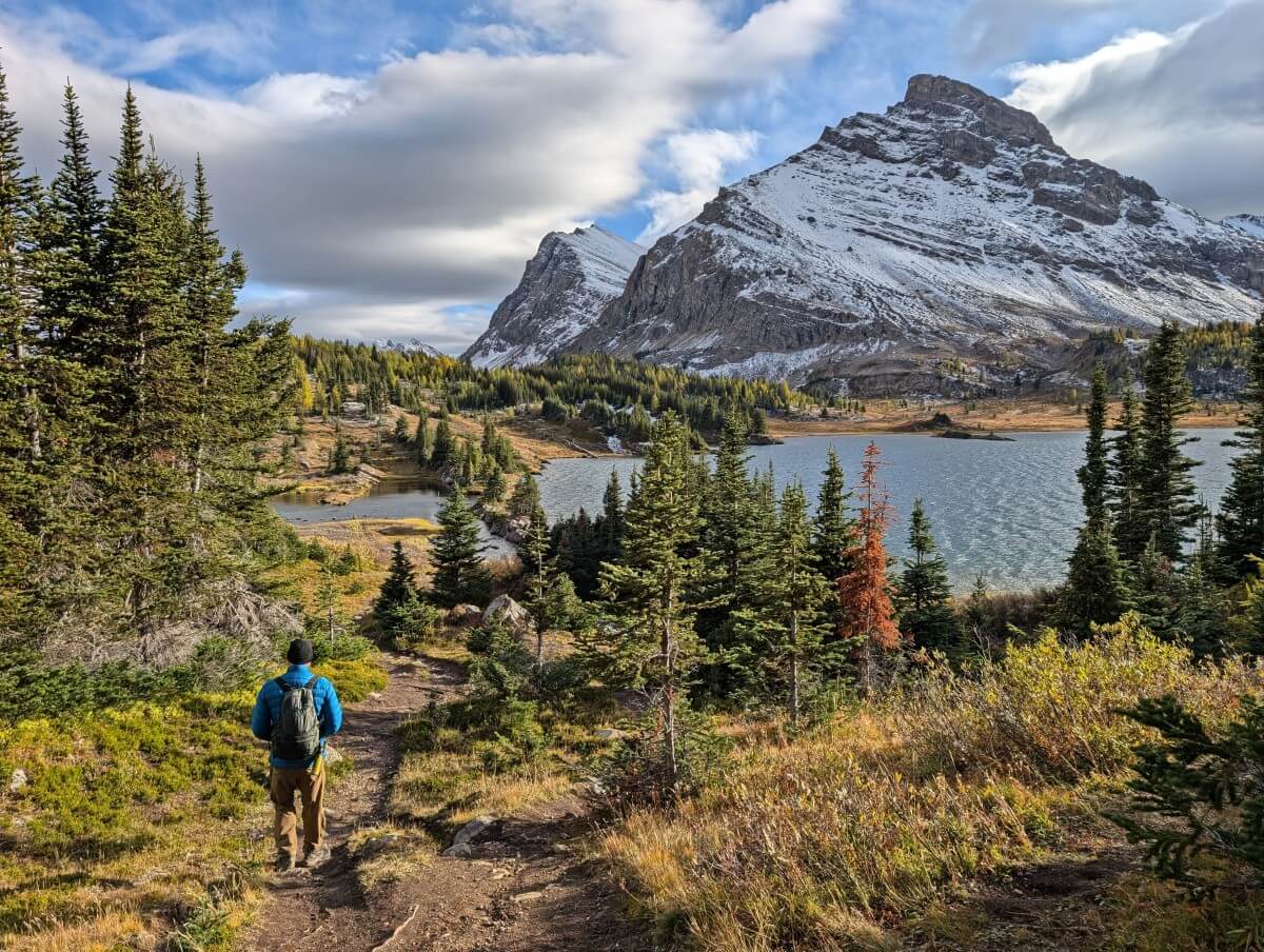 Back view of hiker walking along dirt path towards Baker Lake Campground, with lake in background, backdropped by partially snow covered mountain