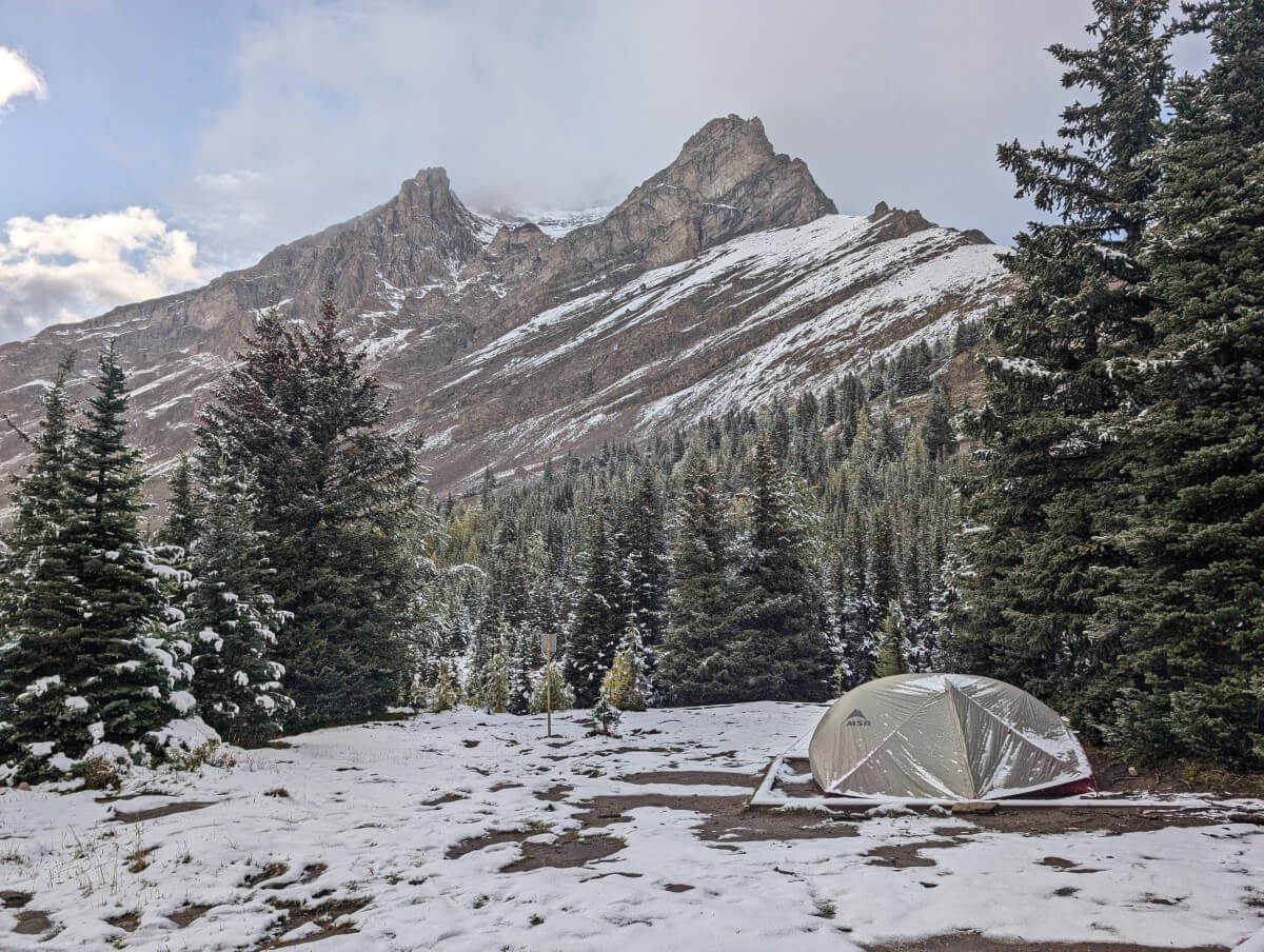 A set up tent sits on a dirt wooded framed tent pad in Baker Lake Campground with forest and mountain backdrop. The landscape is covered in snow. 