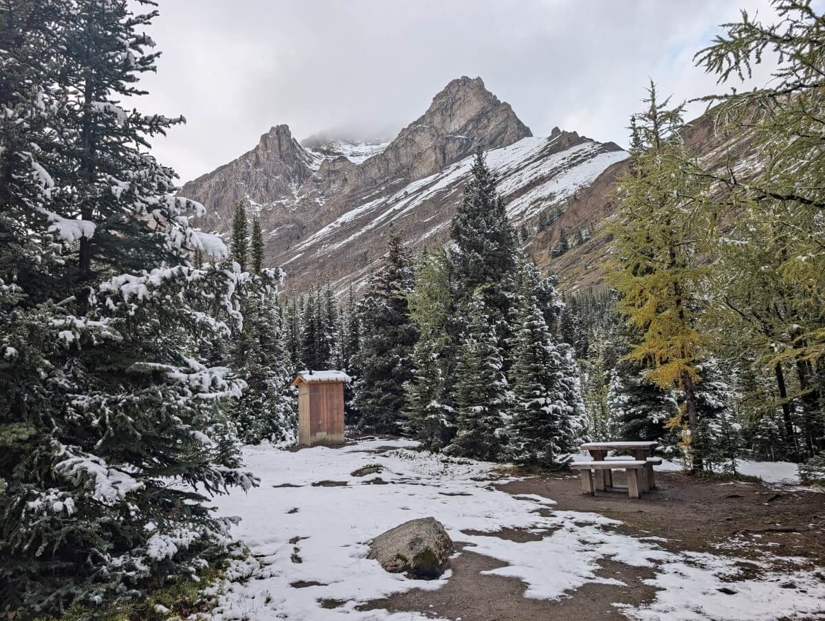 A wooden outhouse and small wooden picnic table in Baker Lake cooking area surrounded by forest and backdropped by mountains. The landscape is partially covered in snow