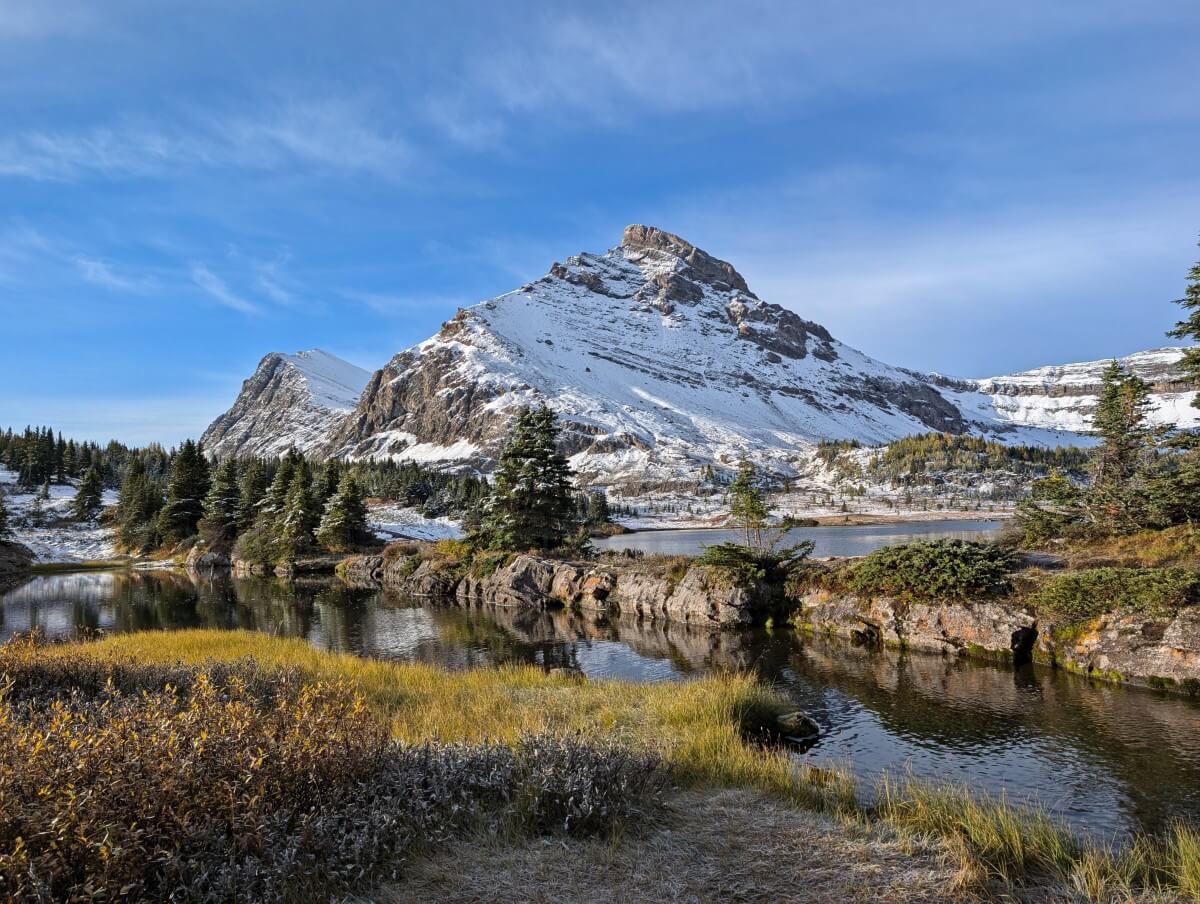 Shore view of Baker Lake, with rock wall separating creek in flow to main part of lake, backdropped by partially snow covered mountain in background