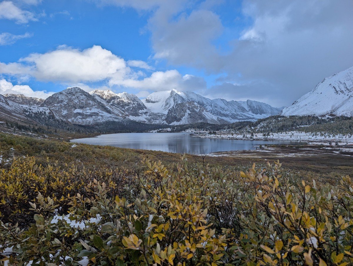 Trail view with autumnal foliage in foreground and Baker Lake in background, backdropped by a range of snow capped mountains