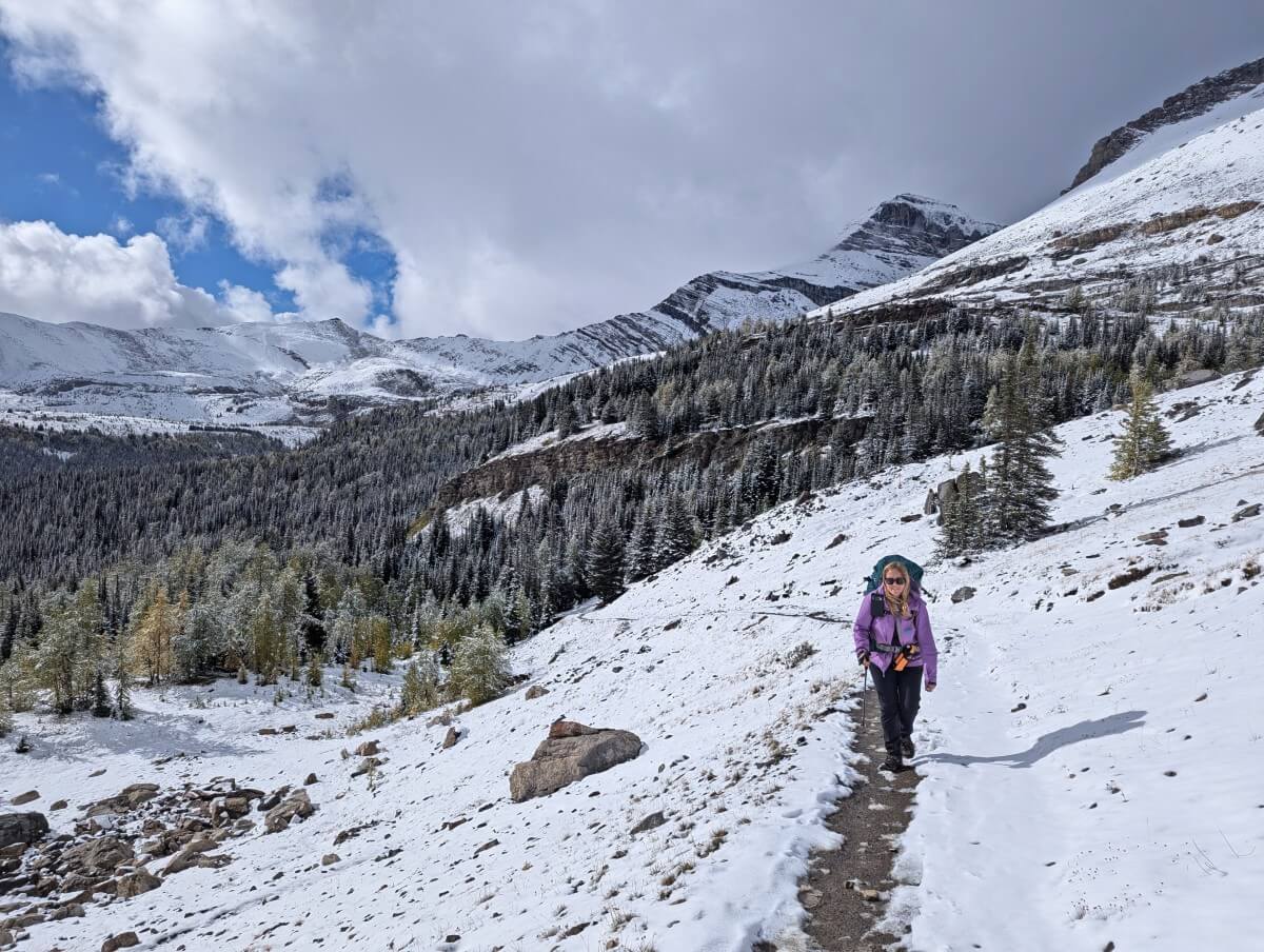 Front view of hiker (Gemma) walking towards camera on dirt trail surrounded by snowy landscape, with snow capped mountains in the background
