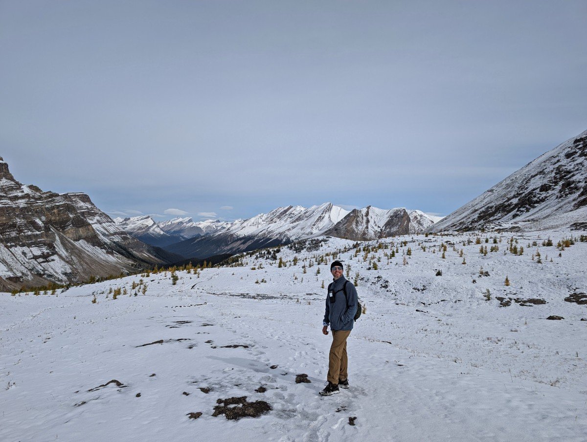 Hiker standing in snow covered landscape on top of Deception Pass, with scattered trees and snow capped mountains behind