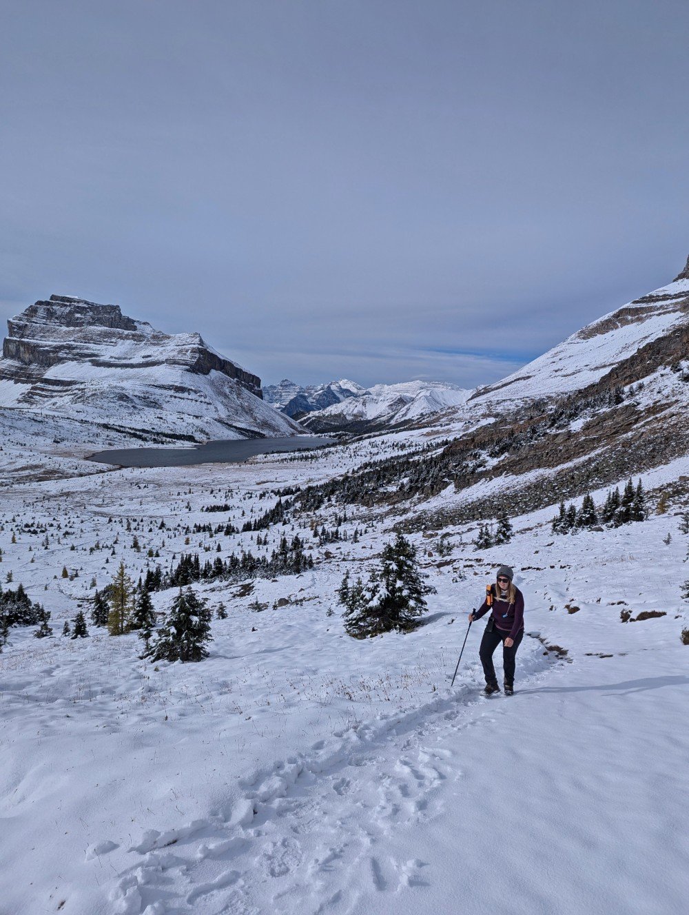 A hiker walks up a snowy trail approaching Deception Pass in Banff National Park. Behind, the open mountainous landscape is snow covered. A large lake is visible