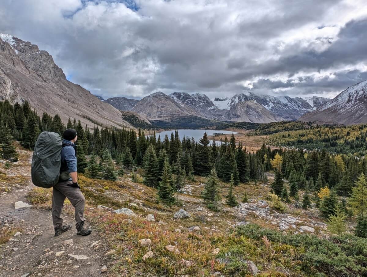 Back view of hiker (JR) with a large backpack standing on dirt trail looking out to lake view below. The lake is surrounded by snowy mountains