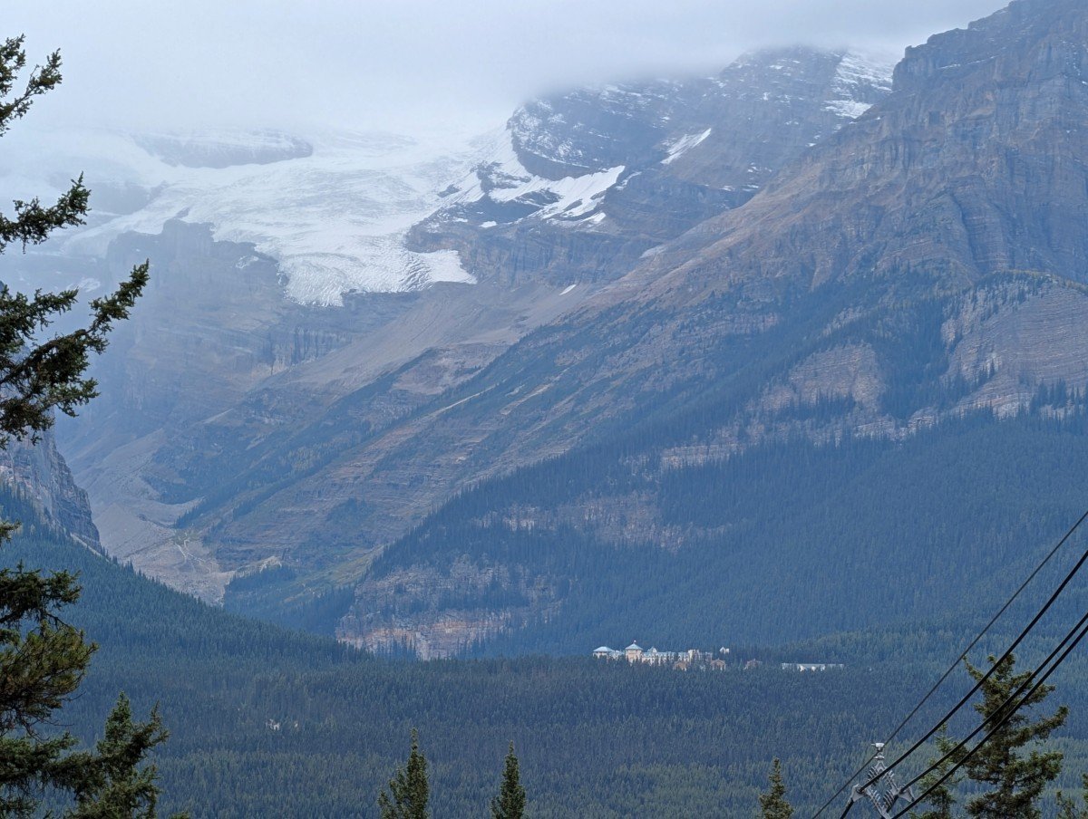 Distant view from the Baker Lake Trail towards the Victoria Glacier (above Lake Louise) and surrounding mountains, with the top of the Fairmont Chateau poking out of the trees