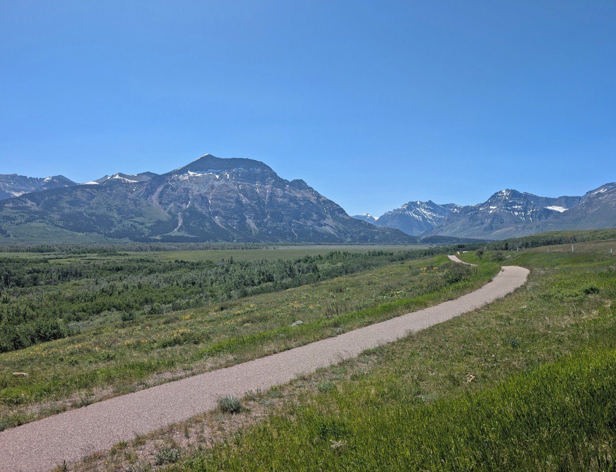 A paved path runs through a prairie grasslands, with views of mountains in the background
