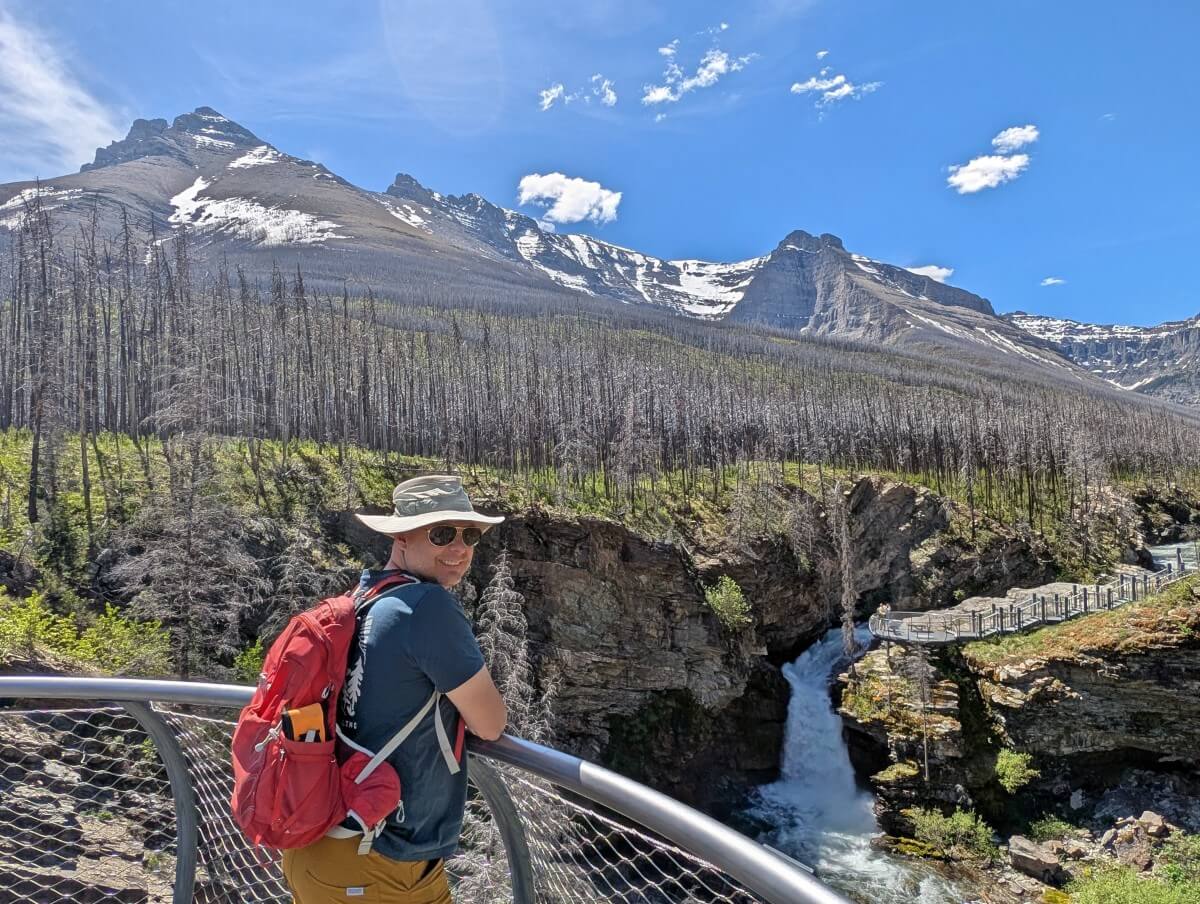 Side view of JR looking back at camera, standing on viewpoint platform in front of Blakiston Falls in Waterton National Park. The backdrop is burned trees with partially snow covered mountains behind