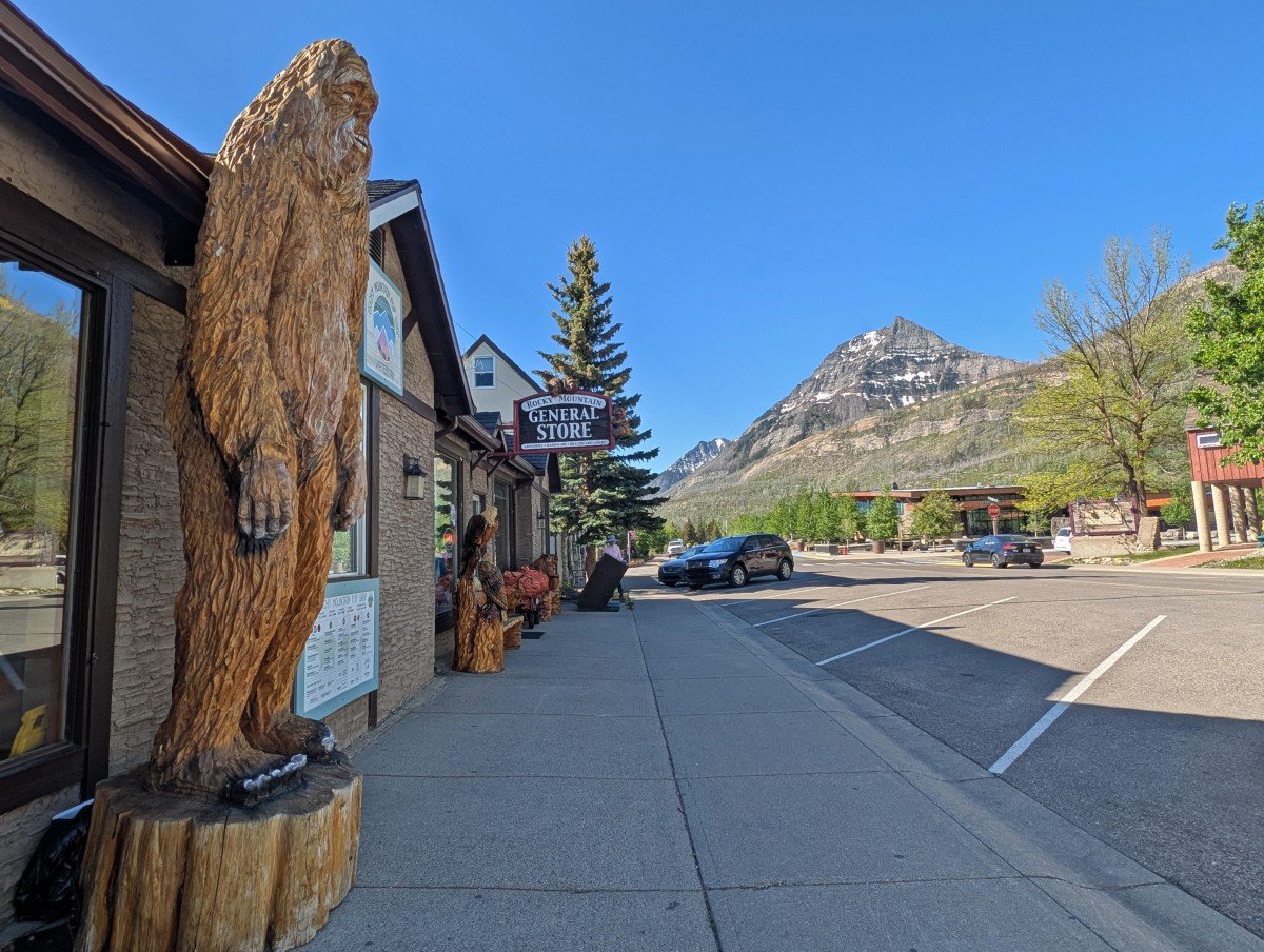 Waterton Townsite street scene with sasquatch carved statue on left, paved path ahead and empty parking spots on right. A mountain is visible in the background