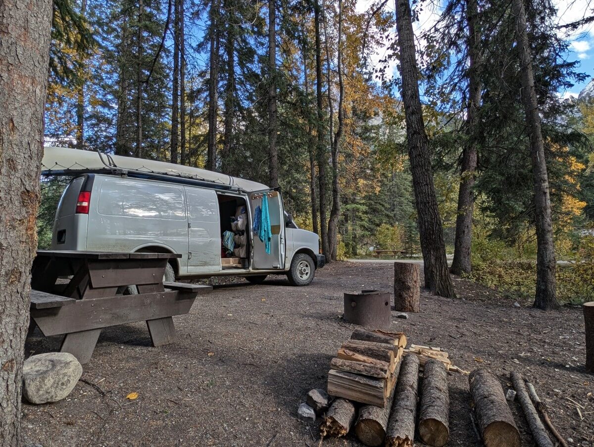 Kicking Horse Campground campsite with parked white van, picnic table, pile of firewood, backdrop of scattered trees