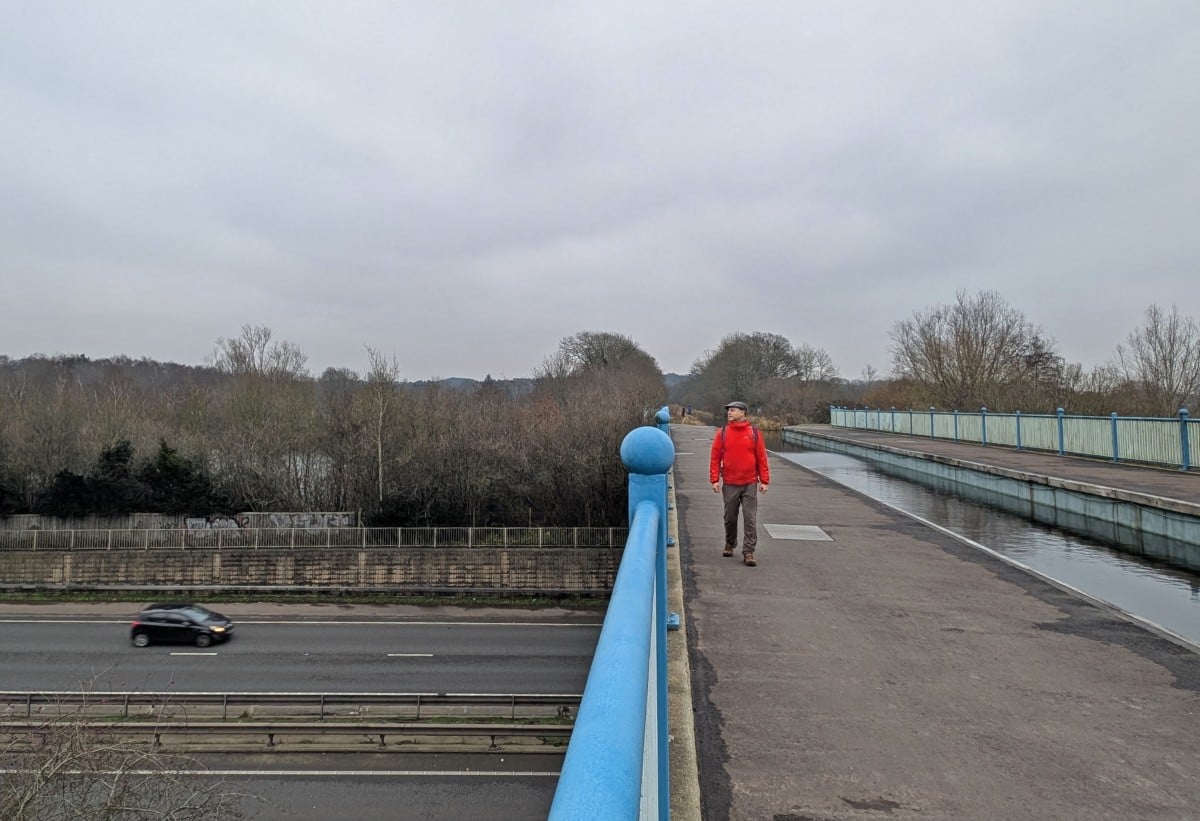 View of man walking towards camera on the concrete towpath next to the Basingstoke Canal on the Ash Aqueduct, which is above a busy road