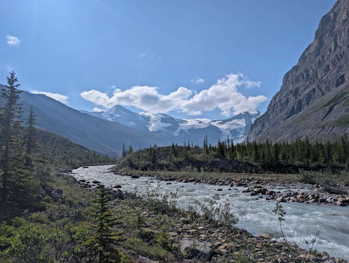 A rushing turquoise coloured river with foliage and small trees either side, snow capped mountains and a glimpse of a glacier in the background
