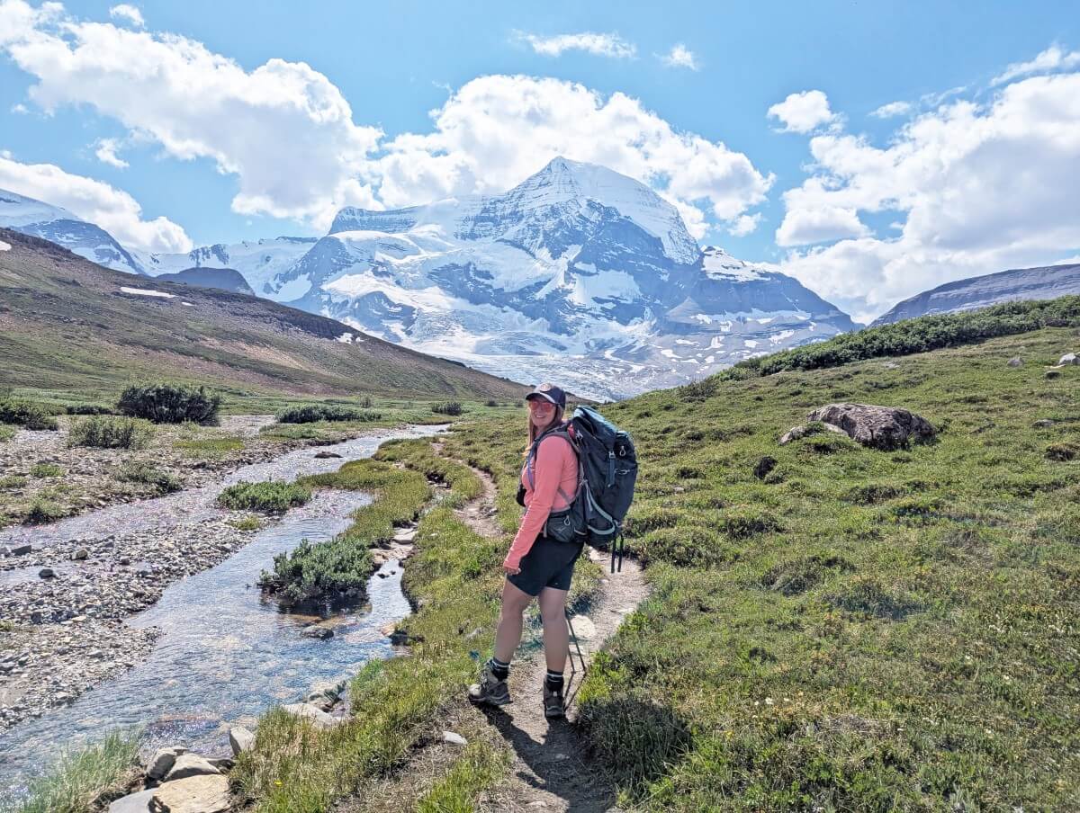 Side view of hiker standing on dirt trail through alpine meadows on Snowbird Pass route, with river on left and large mountain and glacier behind