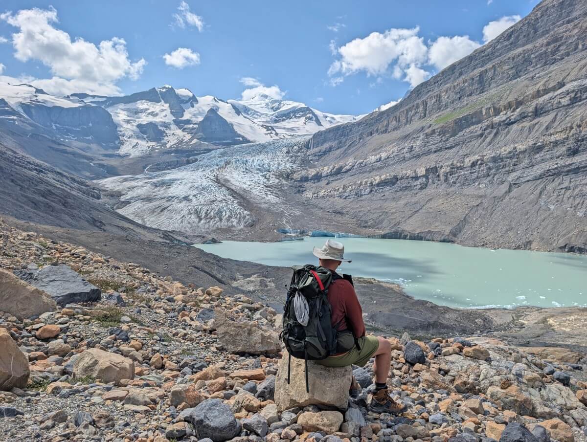 Back view of hiker sitting on rock looking out to view of milky turquoise coloured glacial lake with glacier and snow capped mountains behind