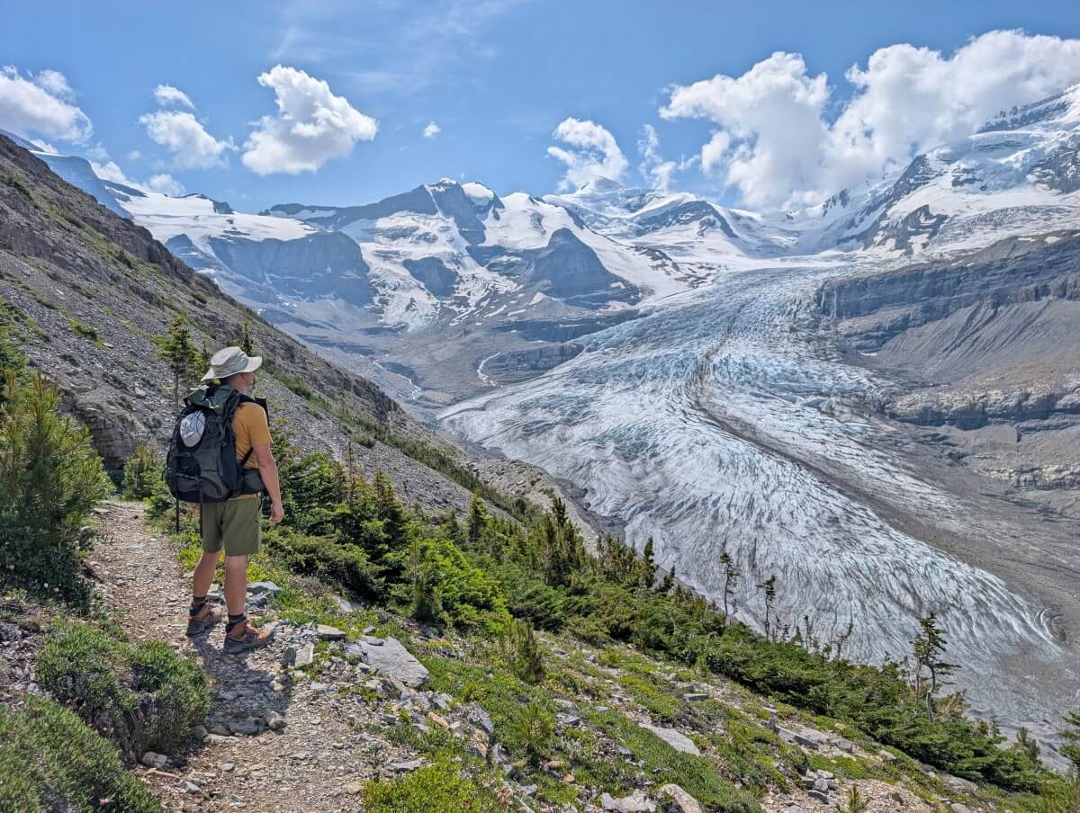 Side view of hiker standing on trail looking out to the huge Robson Glacier, backdropped by snowy mountains