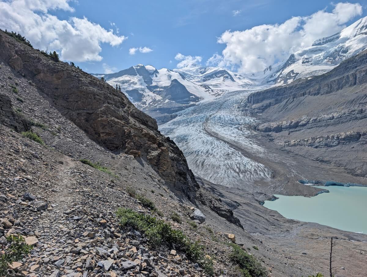 Established trail traversing side of rock slope with view of glacier and glacial lake behind. Some chain fences are visible on the rockface