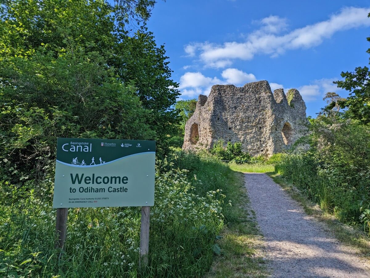 Welcome to Odiham Castle sign next to a path leading to the ruins of a small castle next to the Basingstoke Canal