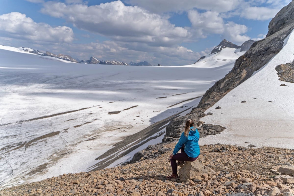 A hiker sits on a rock looking out to the vast Reef Icefield on the Snowbird Pass trail, which stretches endlessly into the distance