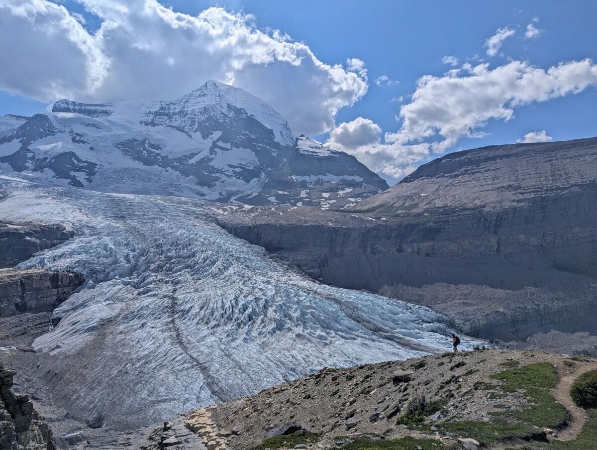 A hiker stands in the distance in front of the huge Robson Glacier, backdropped by Mount Robson