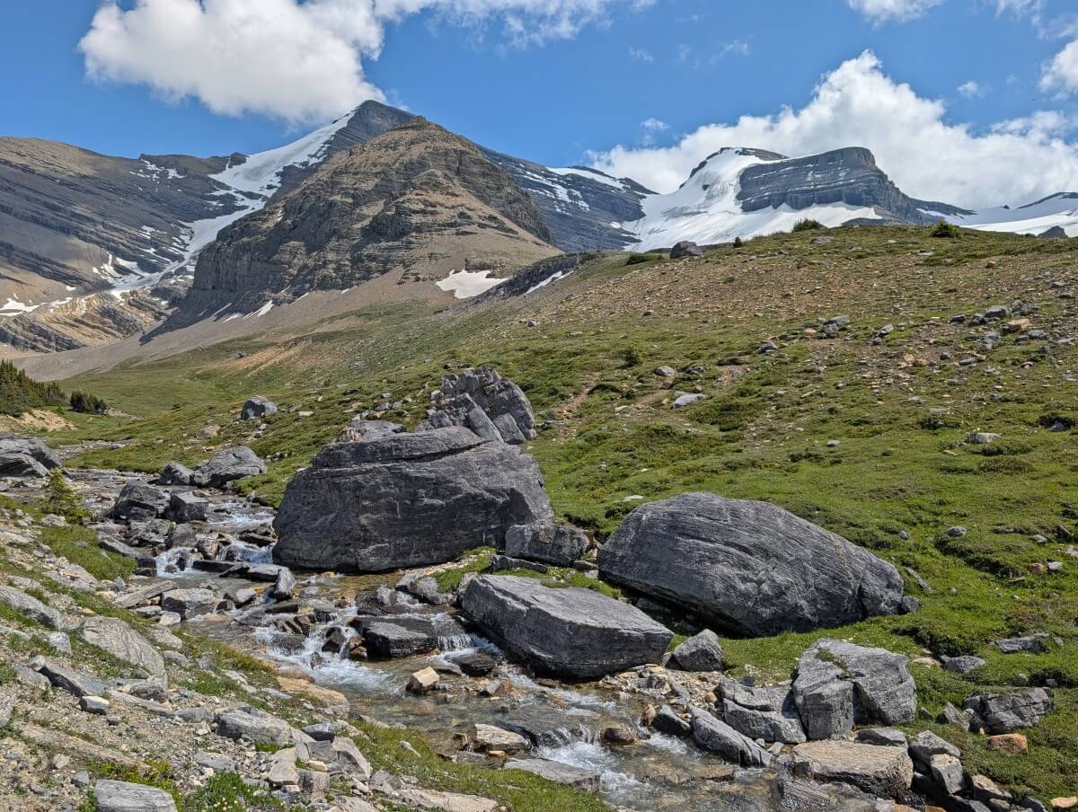 Alpine meadows area on the Snowbird Path trail featuring a fast moving creek next to large boulders, backdropped by meadows and snow capped mountains