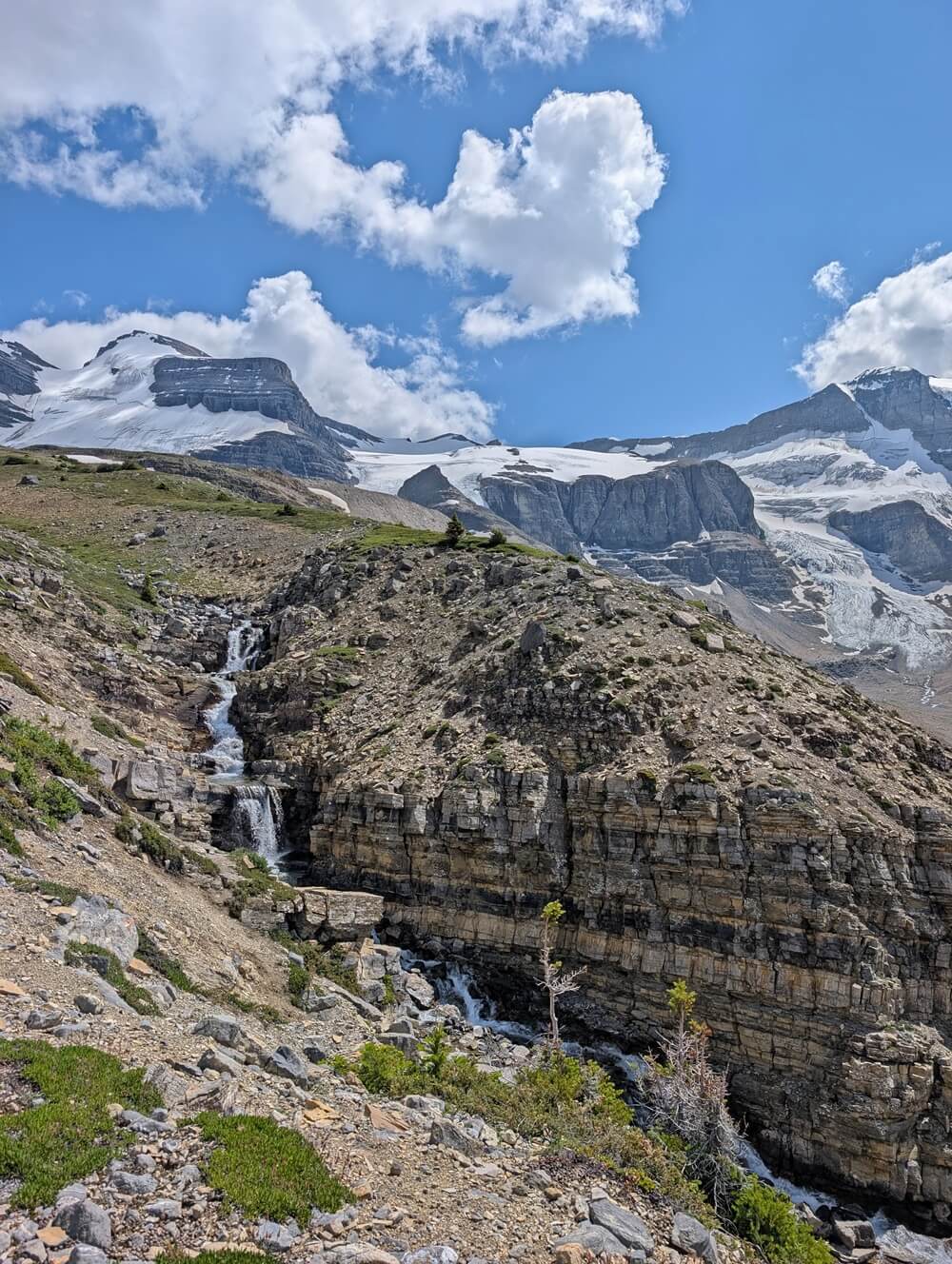 Trail view of rocky area with stream running through, snow capped mountains behind